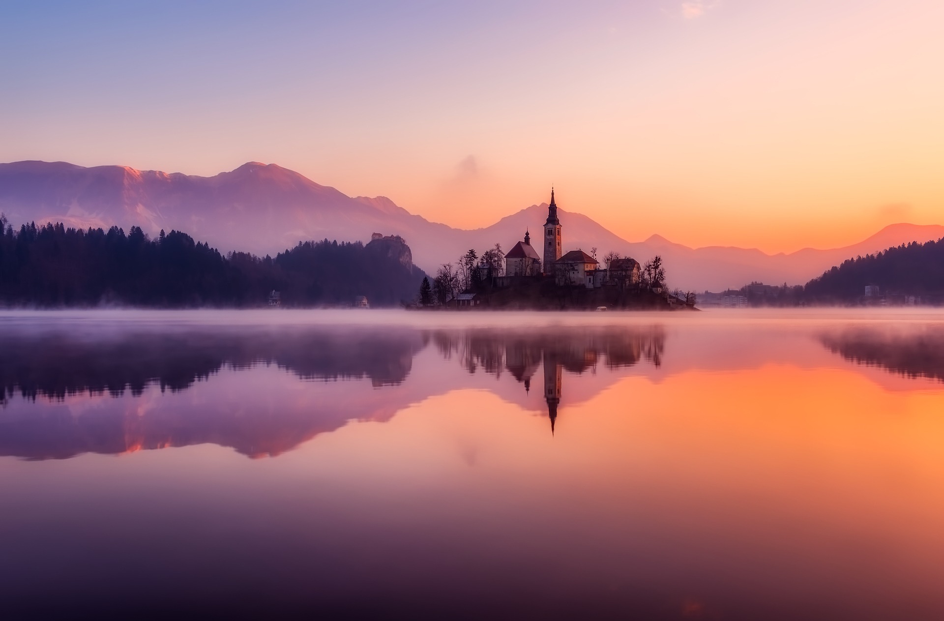 Calm lake at sunrise with a small island church tower and mountains reflected in still water, warm golden and blue tones